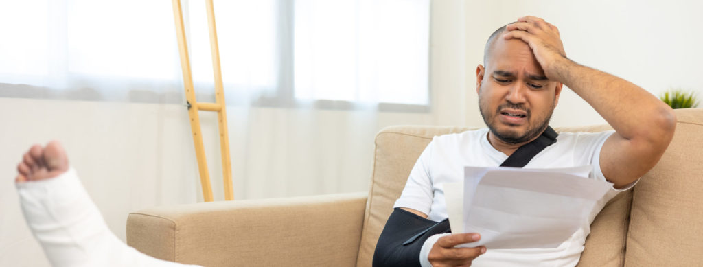 Unhappy, stressed man in cast and arm sling sitting on couch holding medical bill
