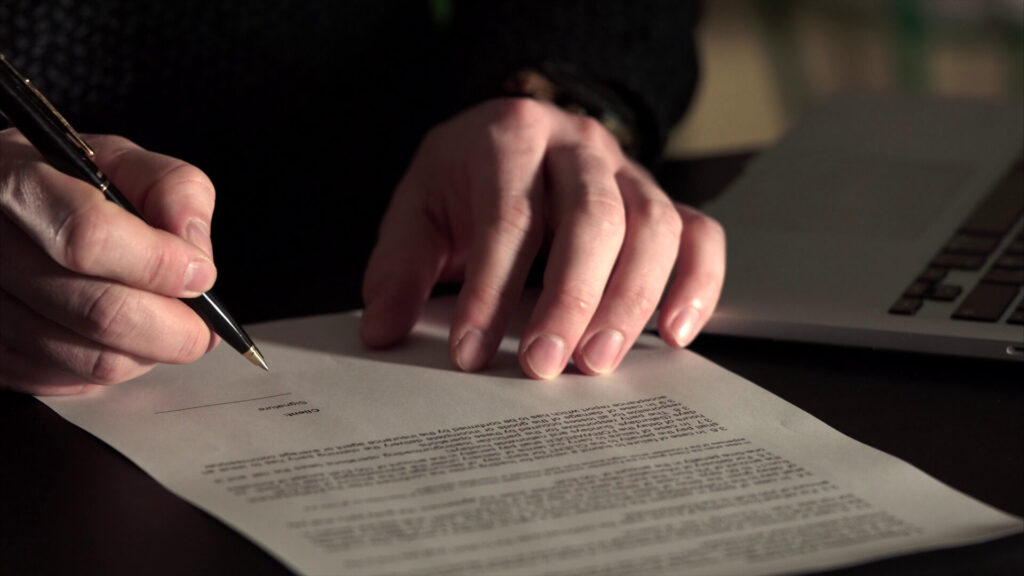 Person signing a legal document with pen next to laptop, representing accident claim settlement.