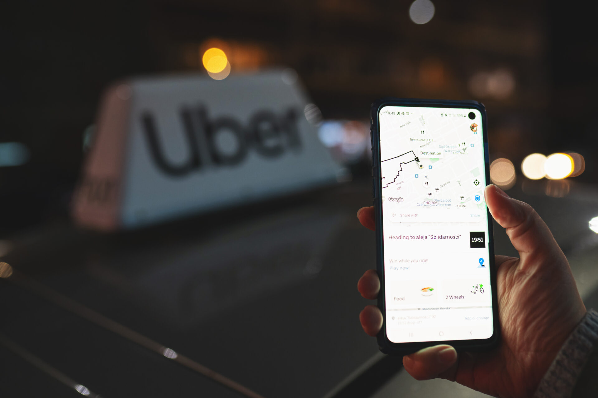 A person holding a smartphone with the Uber app open, showing a ride destination, with an Uber vehicle in the background at night.