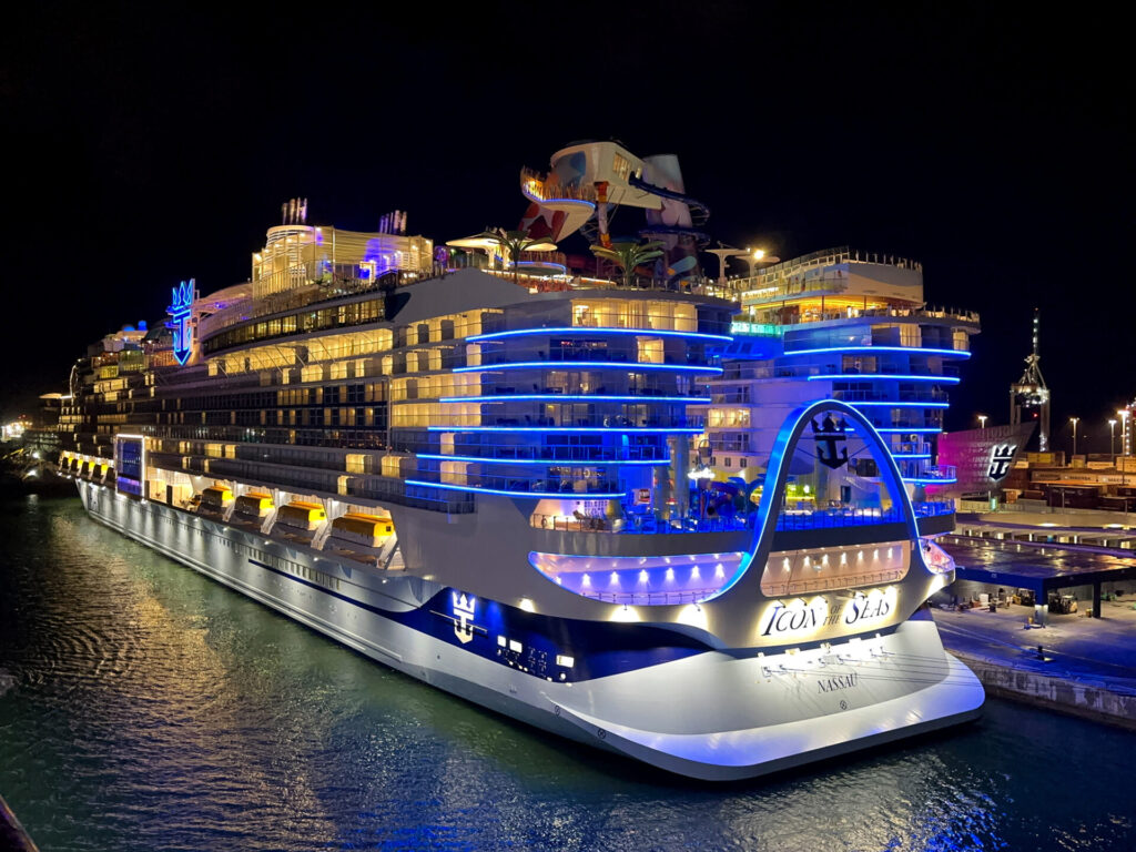 Icon of the Seas, a Royal Caribbean cruise ship, docked at night in Miami with vibrant blue and yellow lights illuminating its decks.