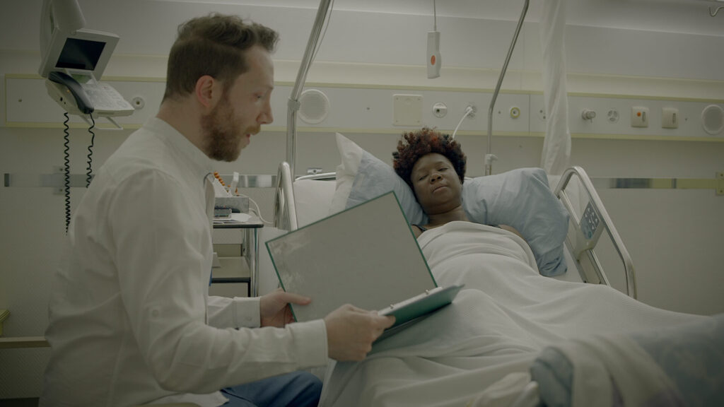 A doctor in a white coat sits beside a hospital bed, holding a clipboard while speaking to a patient who is lying in bed, covered with a white blanket. The patient appears tired and is listening attentively.