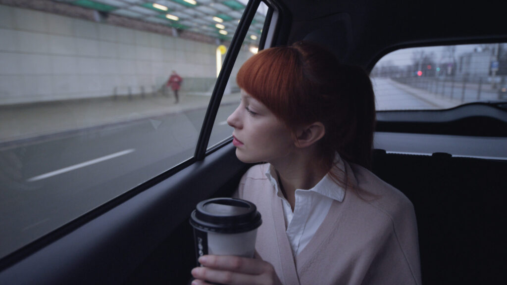 A young woman with red hair sits in the back seat of a car, gazing out the window while holding a takeaway coffee cup. The cityscape outside is blurred, giving a contemplative atmosphere.