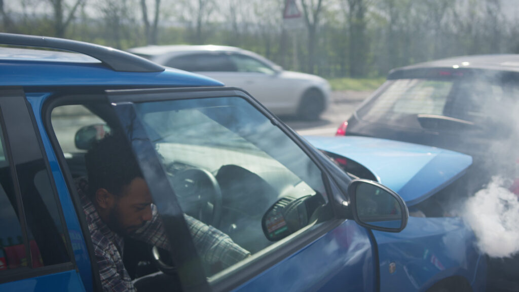 A young male driver sits in his blue car, appearing disoriented after a rear-end collision. Smoke rises from the damaged vehicle as other cars pass by on the road.