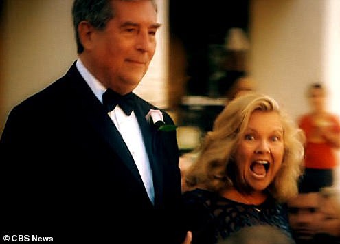 A man in a tuxedo and a woman in a black dress stand together at an event, with the woman appearing emotional and the man looking serious, in a blurred background with other people.