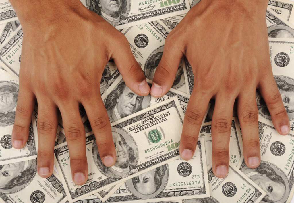 Two hands spread over a pile of U.S. $100 bills, symbolizing wealth and financial compensation.