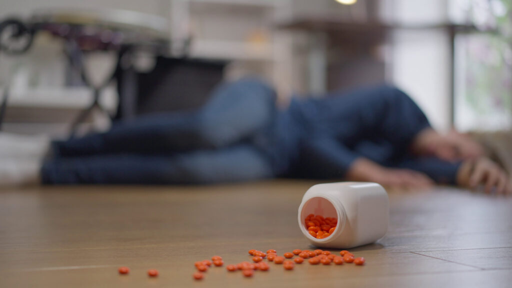 A blurry orange pill bottle sits in the foreground with a person lying on the floor, blurred in the background, suggesting a pharmacy error incident.