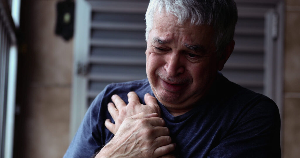 Elderly person in a blue shirt clutching their chest near a window with closed shutters.