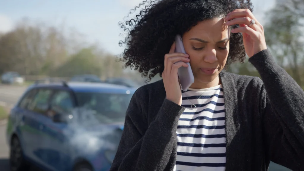 A woman in a striped shirt and cardigan stands on the phone next to a blue car after an accident, with a rural background of trees and a road.