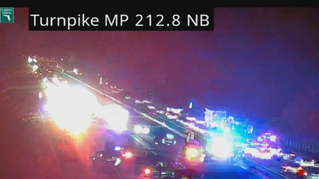 Traffic camera image showing emergency vehicles with flashing lights at the scene of a crash on Florida's Turnpike at mile marker 212.8 northbound, causing a traffic backup at dusk.