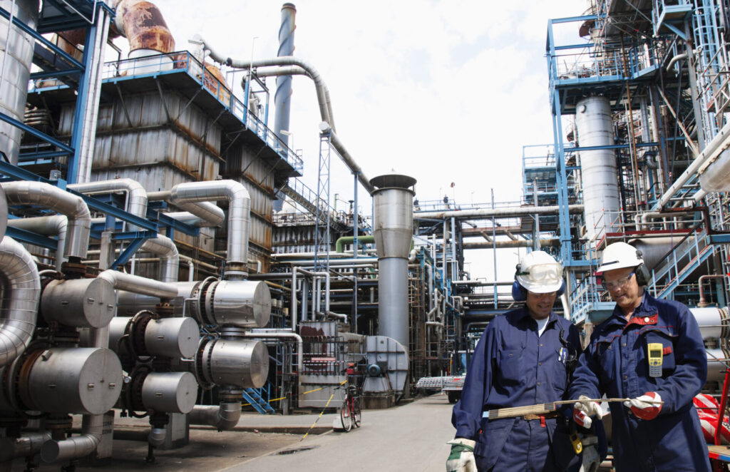 Two workers in hard hats and blue coveralls inspect equipment in an industrial factory setting with large pipes and machinery.