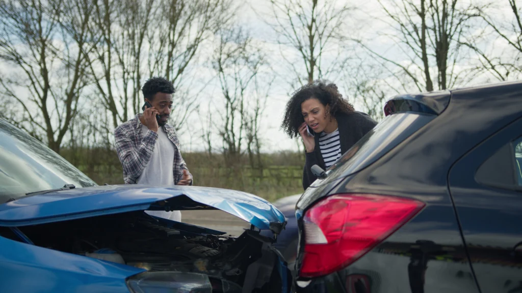 Two individuals stand near a car accident scene with a blue car showing significant front-end damage and a black car with rear damage. Both are on their phones, likely reporting the incident, in a rural setting with bare trees in the background.
