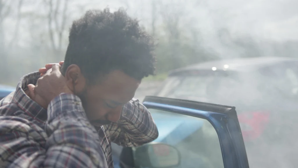 A person in a plaid shirt stands next to a car with a damaged rear, holding their neck, suggesting possible injury after an accident. Smoke or haze is visible in the background, indicating a recent crash.