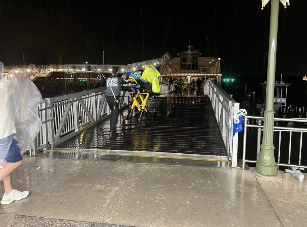 Emergency responders wheel an injured person on a stretcher at the rainy Disney Magic Kingdom ferry boat crash scene at night, with the damaged dock and boat in the background.