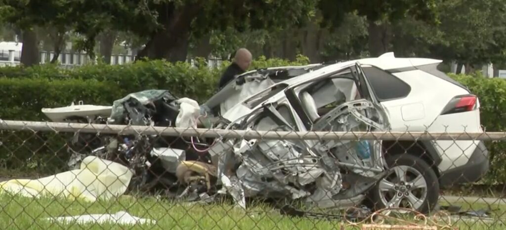 Crime scene investigator examining wreckage of a white SUV in a fatal three-vehicle car accident in North Miami Beach, with yellow police tape and debris scattered on grass.