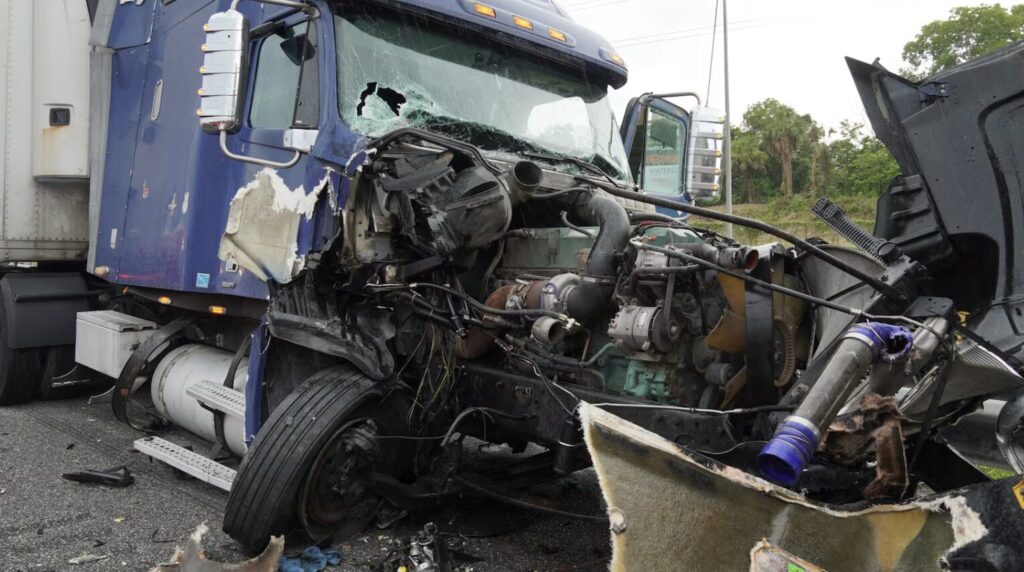 Damaged blue semi-truck after a crash, with a shattered windshield, exposed engine, and scattered debris on the road.
