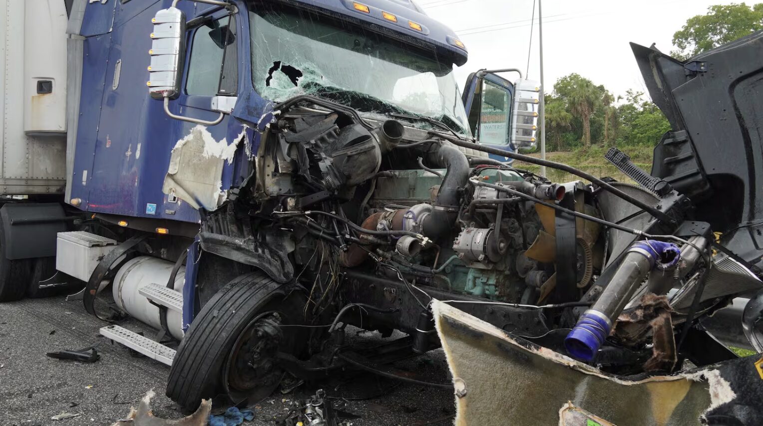 Damaged blue semi-truck after a crash, with a shattered windshield, exposed engine, and scattered debris on the road.