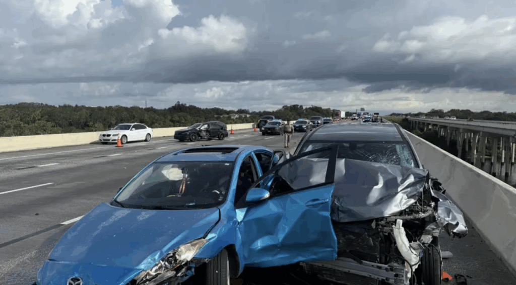 Scene of a multi-vehicle crash on I-75 bridge over Alafia River, showing damaged blue and gray cars, police presence, and traffic cones in Gibsonton, Florida.