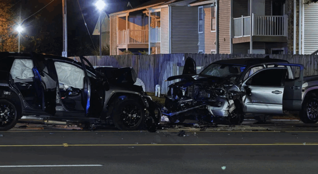 Nighttime scene of a multi-vehicle car accident on 9th Avenue in Pensacola, Florida, showing three damaged vehicles—including an SUV, a sedan, and a pickup truck—with doors open, debris scattered on the road, and residential houses in the background.