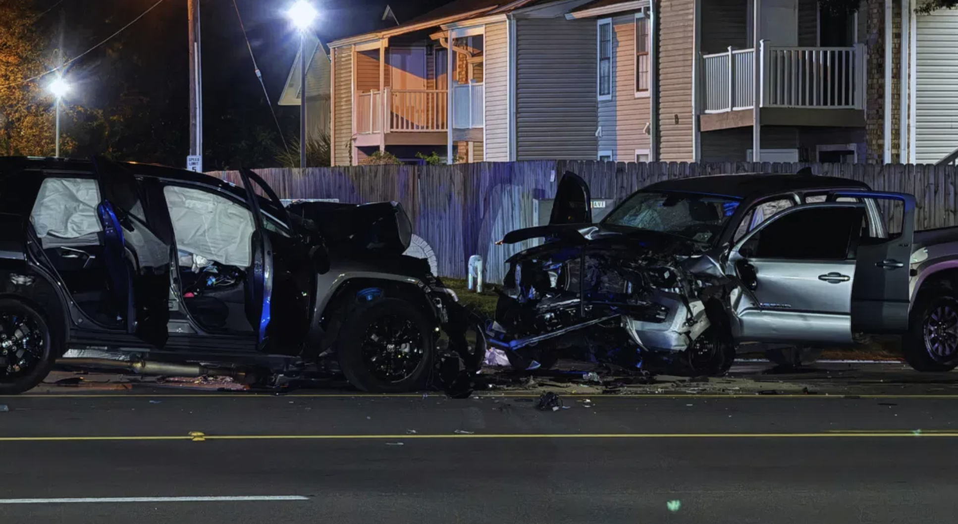 Nighttime scene of a multi-vehicle car accident on 9th Avenue in Pensacola, Florida, showing three damaged vehicles—including an SUV, a sedan, and a pickup truck—with doors open, debris scattered on the road, and residential houses in the background.