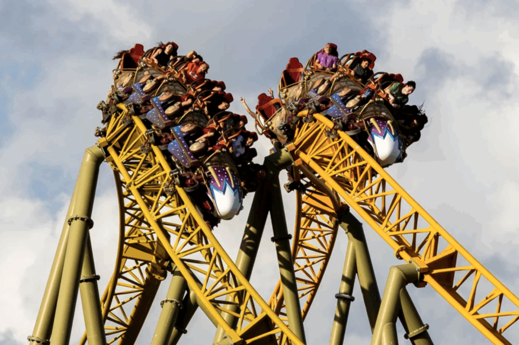 Thrilling action shot of the Stardust Racers dueling roller coaster at Universal's Epic Universe, showing two yellow trains with excited riders in purple and red seats crossing paths at 133 feet high under a cloudy sky, emphasizing the intense ride that recently led to a tragic accident.