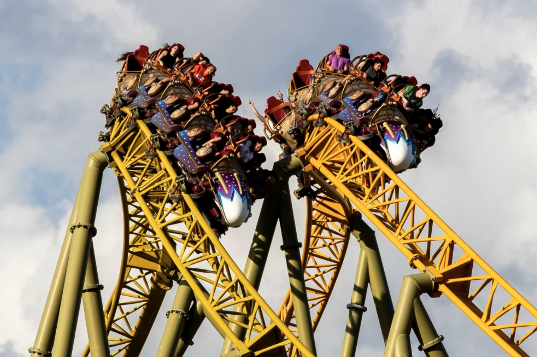 Thrilling action shot of the Stardust Racers dueling roller coaster at Universal's Epic Universe, showing two yellow trains with excited riders in purple and red seats crossing paths at 133 feet high under a cloudy sky, emphasizing the intense ride that recently led to a tragic accident.