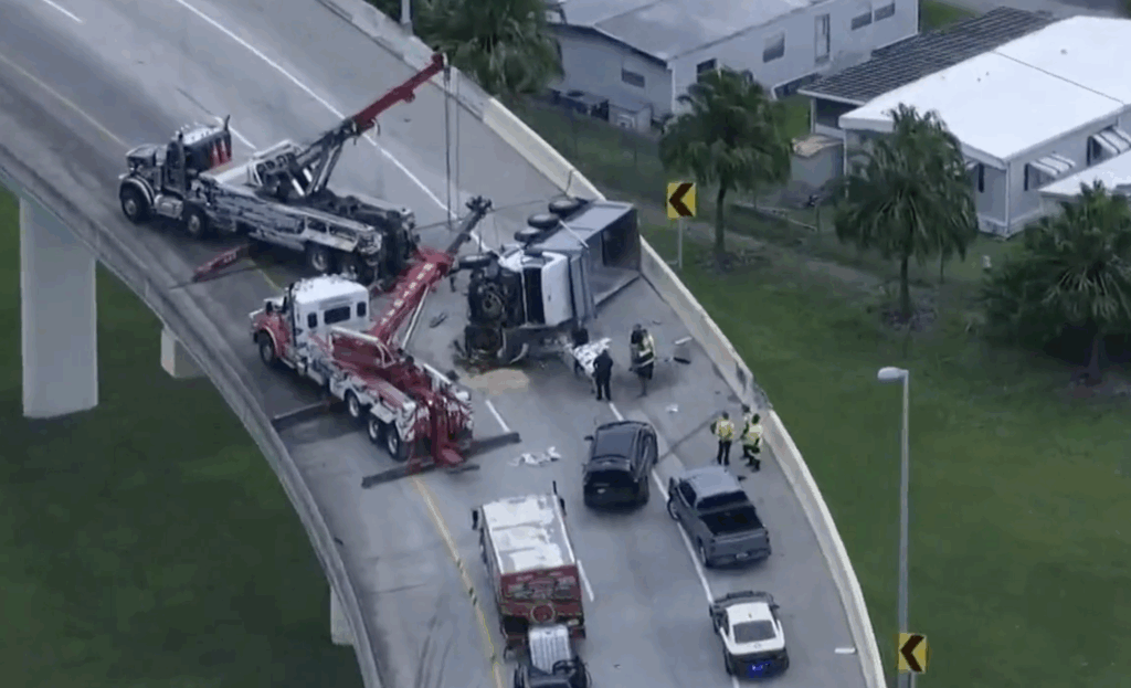 Aerial view of a truck rollover crash on a highway with tow trucks and emergency responders working on the scene.