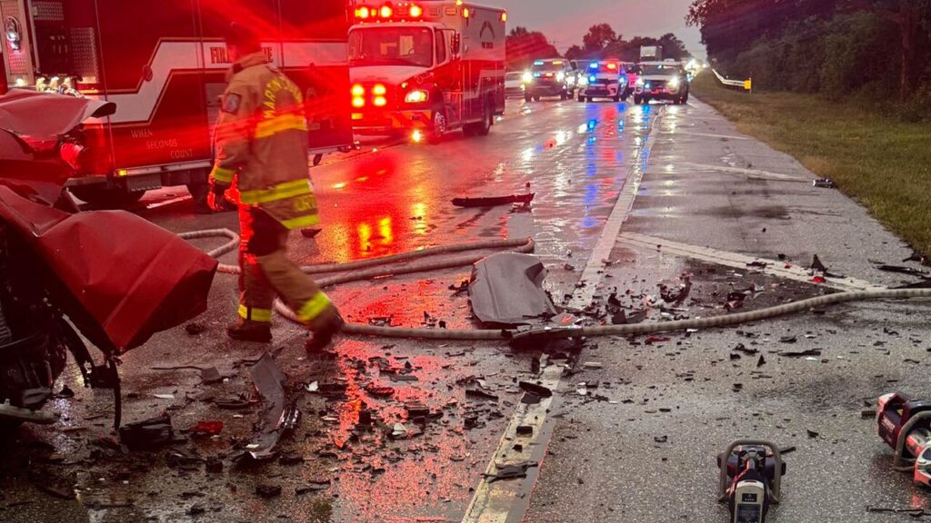 A firefighter stands amid wreckage on a wet State Road 710 after a fatal car accident, with emergency vehicles and debris scattered.