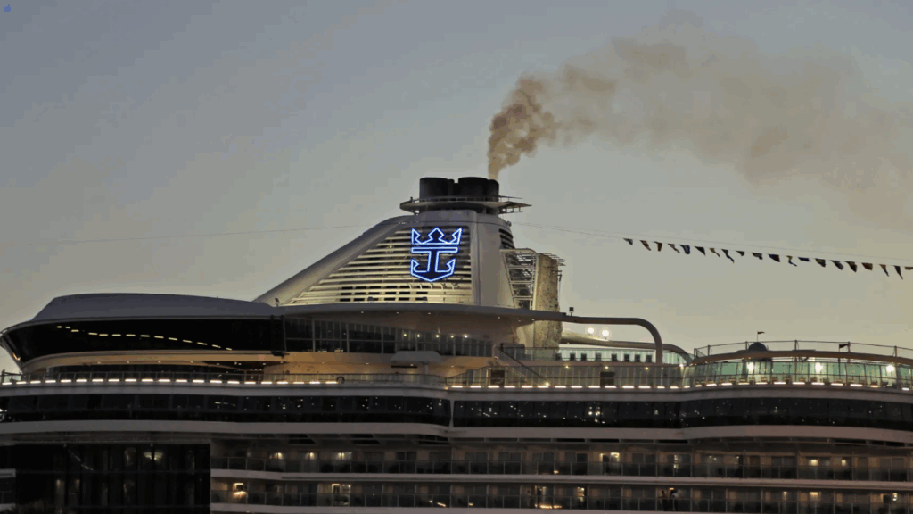 A large cruise ship with a prominent logo emitting smoke from its chimneys at dusk, with multiple decks and festive flags.