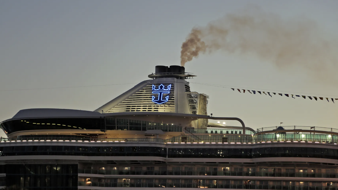 A large cruise ship with a prominent logo emitting smoke from its chimneys at dusk, with multiple decks and festive flags.
