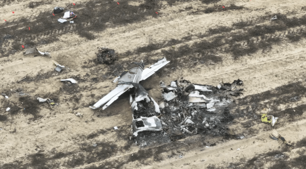 Aerial view of a crashed airplane wreckage in a field with scattered debris and emergency markers.