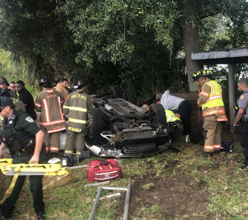 Emergency responders and firefighters working to free a trapped person from an overturned car at a multi-vehicle crash scene in Holly Hill, with medical equipment and police present.