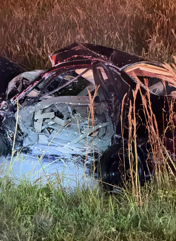 Severe car accident wreckage in a grassy field at night showing extensive side-impact and roof damage.