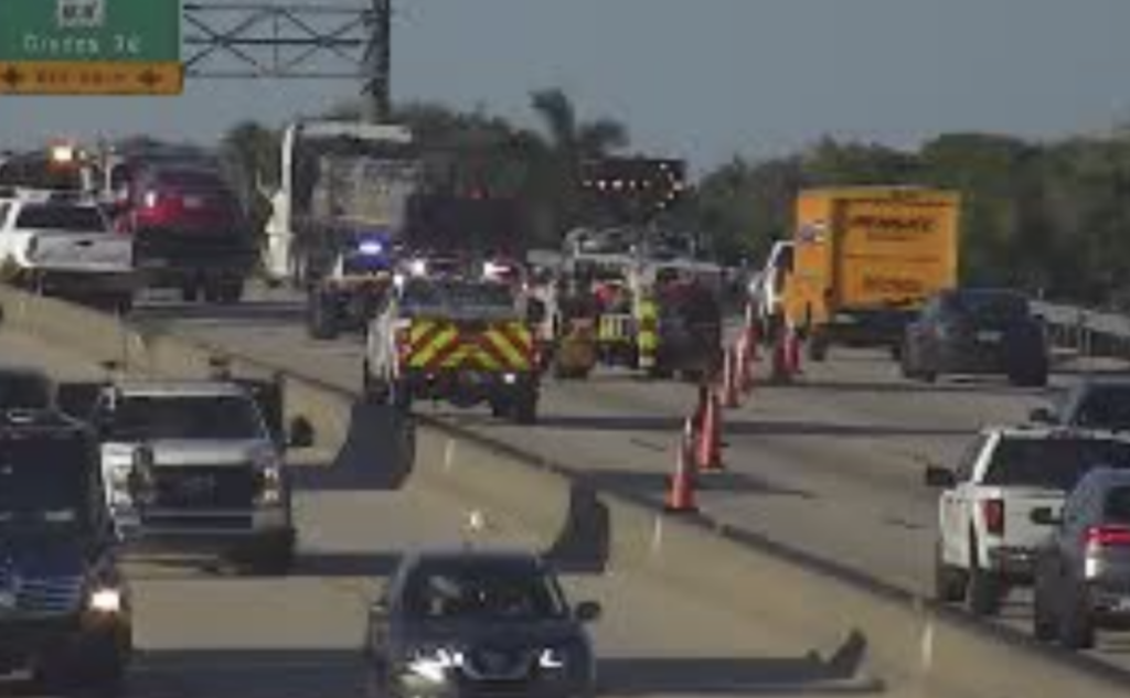 Traffic camera view of a multi-vehicle accident scene on the Florida Turnpike in Boca Raton showing fire rescue vehicles with flashing lights, orange traffic cones diverting lanes, a yellow commercial box truck, and backed-up traffic in both directions under a highway sign for Glades Road exit.